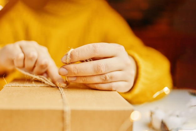 주황색 스웨터를 입은 소녀는 휴일 선물 상자 포장에 끈을 묶습니다.