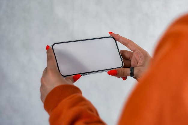 La ragazza con un maglione arancione con belle unghie tiene in mano un modello di smartphone con schermo bianco. tecnologia di simulazione.