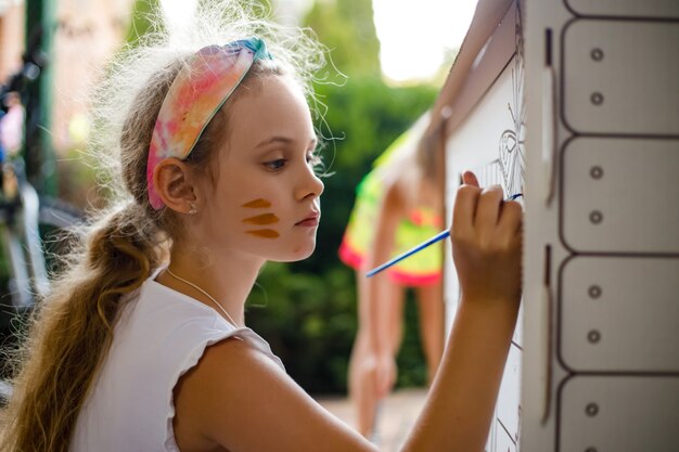 Girl paints a cardboard house at summer day, outdoor.