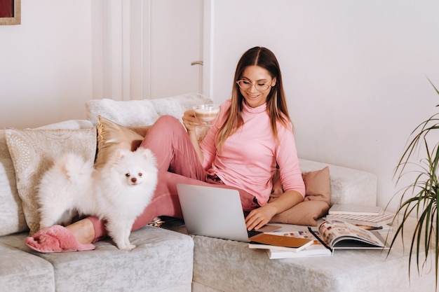 A girl in pajamas at home is working on a laptop with her dog