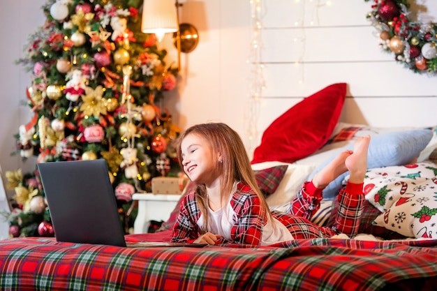 Girl in pajamas lies on a bed on red plaid of a Christmas tree and looks at the monitor of the laptop.