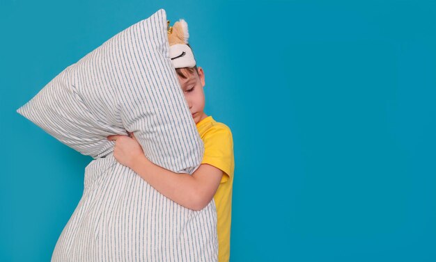 Girl in pajamas and with an eye mask stands on a blue background with a pillow