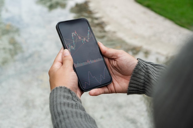 Girl in the park holding a smartphone with Financial stock market graph on the screen Green area and an artificial lake Stock Exchange