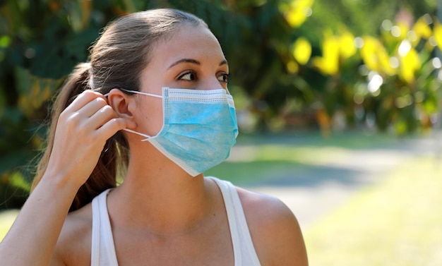 Girl in the park puts on a medical mask. Young sporty woman wearing surgical mask in city park. Copy space.