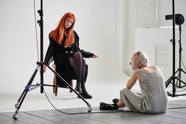 Девушка-фотограф фотографирует фотомодель в черном, сидя на стуле на белом фоне в студии