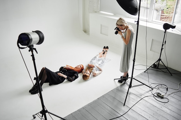 Girl photographer photographing fashion models in a clear coat with a pomegranate mask on the face and black clothes lying on white floor in Studio