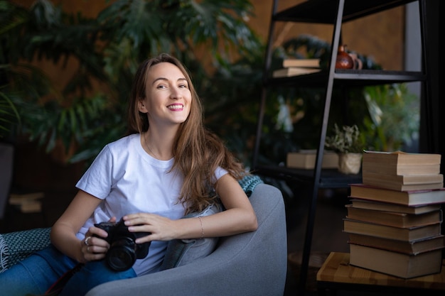 A girl photographer with a camera smiles while sitting in a chair with many books