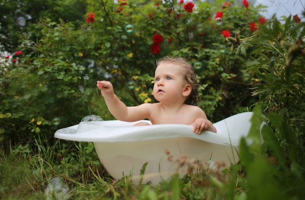 A girl in a picturesque garden in a small white bath catches soap bubbles among the red flowers