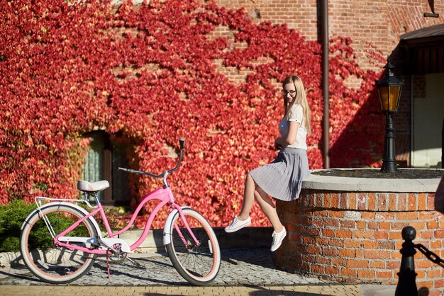 Girl at pink bicycle on sunny day near wall overgrown with red ivy