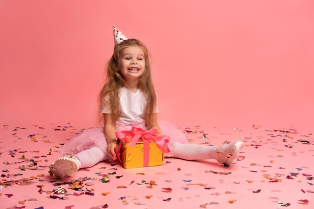 A girl in a pink cap and pink tutu sits on a pink background with confetti on the floor and smiles.