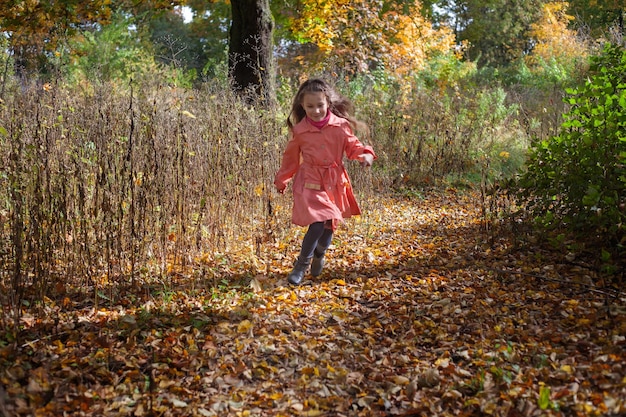 ピンクのマントを着た女の子が葉の上で秋に公園を走る