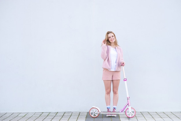 Girl in pink clothes stands with a scooter on the background of a white wall, looks at the camera and smiles