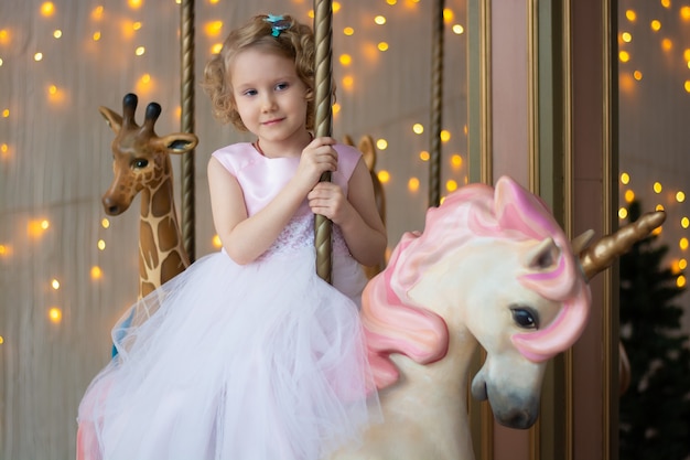 Photo girl in a pink dress and a crown on a carousel with a white pony