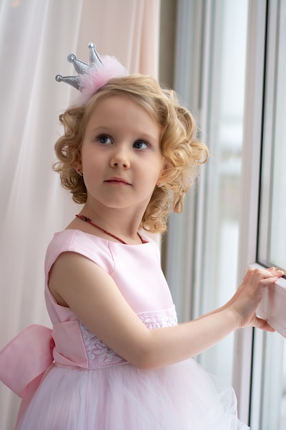 girl in a pink dress and a crown smiles near the window