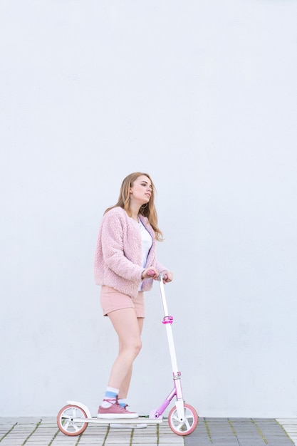 Girl in a pink fur coat and shorts stands with a kick scooter on the background of a white wall