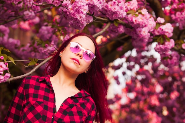 Girl in pink glasses with long pink blowing hair. Spring or summer beauty teen girl under the blooming sakura flowers. Fantasy Magical Spring Garden and dreamer woman