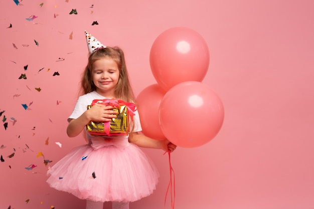 A girl in a pink party hat holds a gift box and smiles.