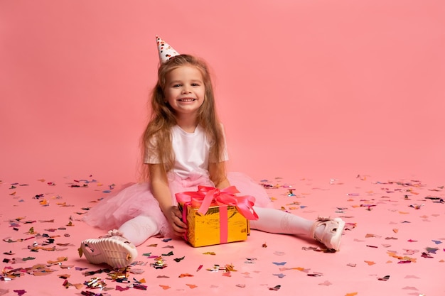 A girl in a pink party hat sits on a pink background with confetti on the floor.
