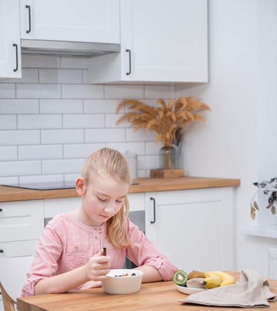 A girl in a pink shirt is sitting at the kitchen table and eating yogurt with granola and fresh fruit The concept of healthy eating