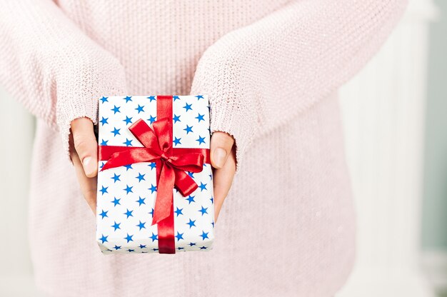 A girl in a pink sweater, holds in her hands a gift with blue stars and a red ribbon. On a light background. Concept on the theme of holidays.