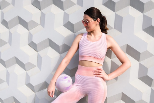 A girl in a pink tracksuit stands with a training ball near the wall