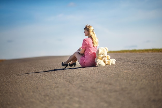 Girl in plaid suit sits on asphalt road with teddy bear concept of loneliness waiting for happiness