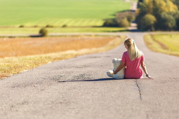 Девушка в клетчатом костюме сидит на асфальтированной дороге с концепцией плюшевого мишки одиночества в ожидании счастья