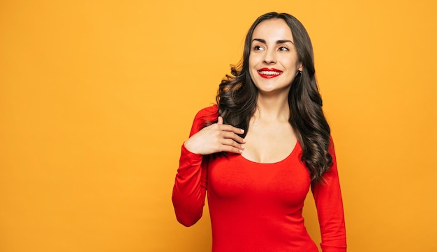 A girl in a playful and pleased mood next to the orange background clothed in bright red sweater is showing her perfect white smile and emotions