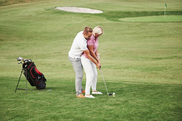 Girl playing golf and hitting by putter on green. Her teacher helps to explore the technique and make her first strikes