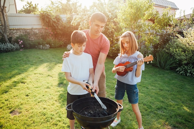 Girl playing ukulele standing near her father and brother cooking food on barbecue