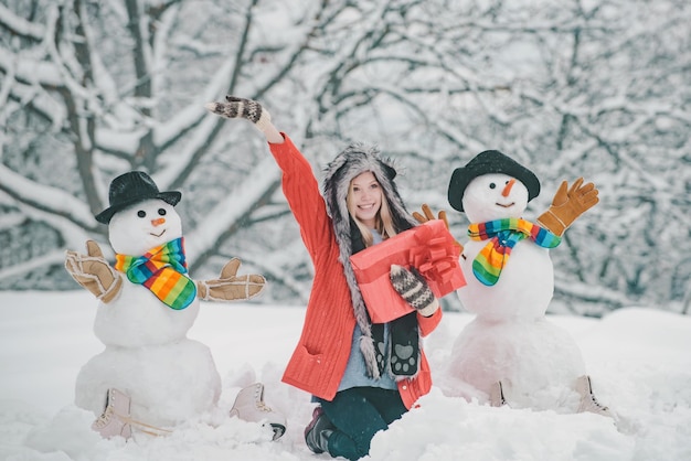 Ragazza che gioca con il pupazzo di neve nel parco invernale pupazzo di neve e giovane donna divertente con cappello e sciarpa invernali