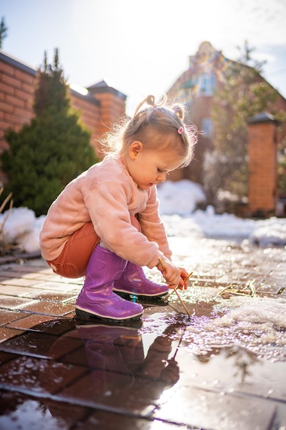 Girl plays in a puddle with wooden stick in spring day at sunlight