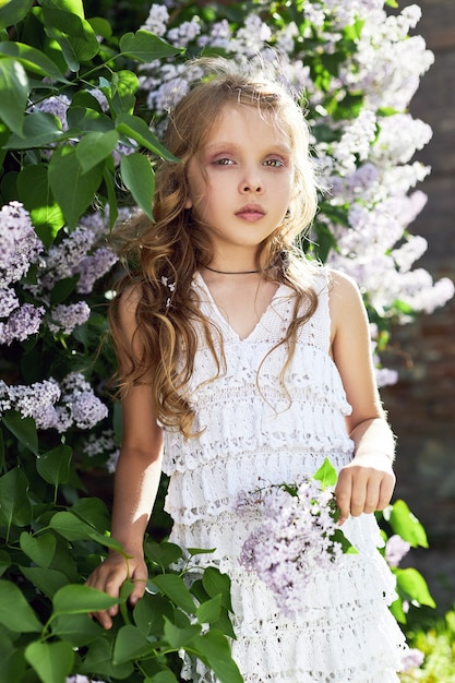 Girl poses in a lilac bush in the spring. Romantic portrait of a child in flowers in the sun light