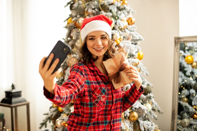 The girl poses and takes a selfie near the Christmas tree. A woman congratulates a relative online by phone. She is holding a gift in her hand and smiling.