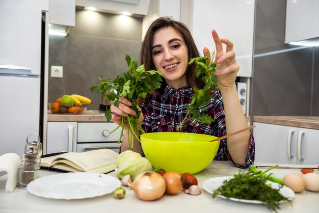 健康的なライフスタイルのためにさまざまな野菜と緑のサラダを準備しながら、台所のテーブルでポーズをとる女の子