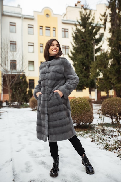 Girl posing on road on winter background Glamorous funny young woman with smile wearing stylish grey long fur coat Fur and fashion concept Beautiful people