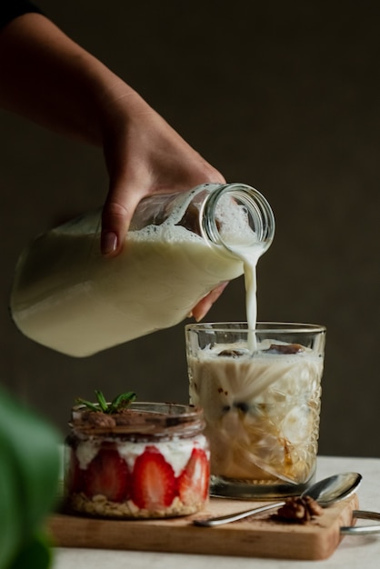 A girl pours milk into a glass with frozen coffee next to tirramisu with strawberries