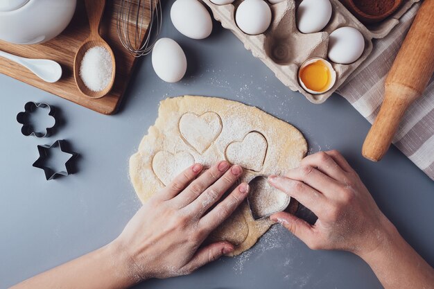 La ragazza prepara i biscotti a forma di cuore, composizione piatta su uno sfondo grigio. tagliabiscotti e pasta nelle mani delle donne. concetto di cibo per san valentino, festa del papà, festa della mamma.