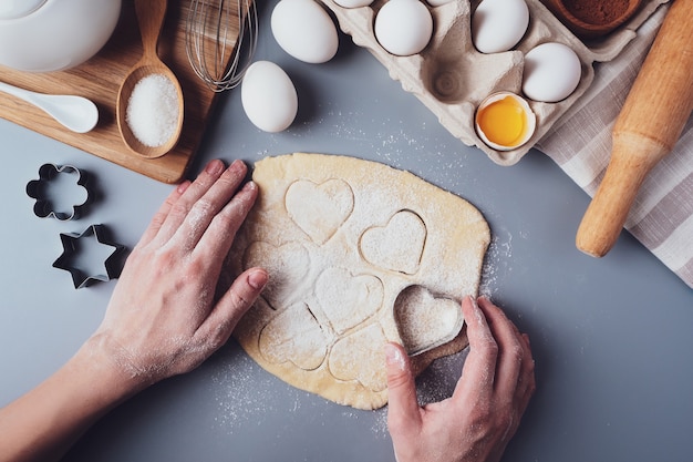 Girl prepares cookies in the shape of a heart, flat lay composition on a gray background.