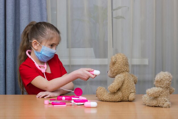 おもちゃで病院で遊んでいる医療マスクの就学前の女の子はテディにタブレットを与えます