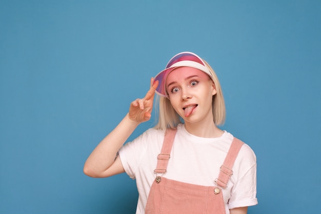 girl in a pretty casual dress, wearing a pink cap and making a funny face on blue.