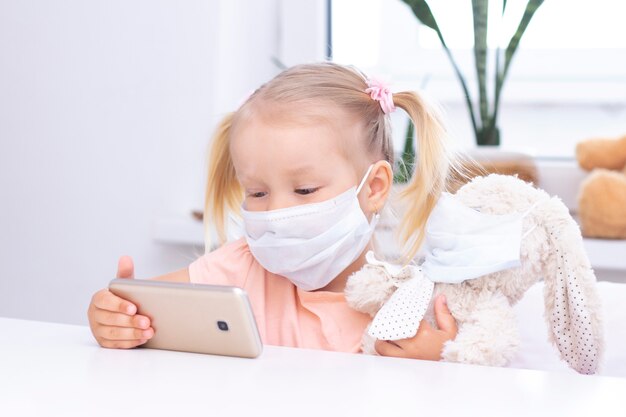 Girl in a protective mask with a toy bunny using a mobile\
phone, a smartphone for video calls, talks with relatives, a girl\
sits at home, online computer webcam, making a video call.