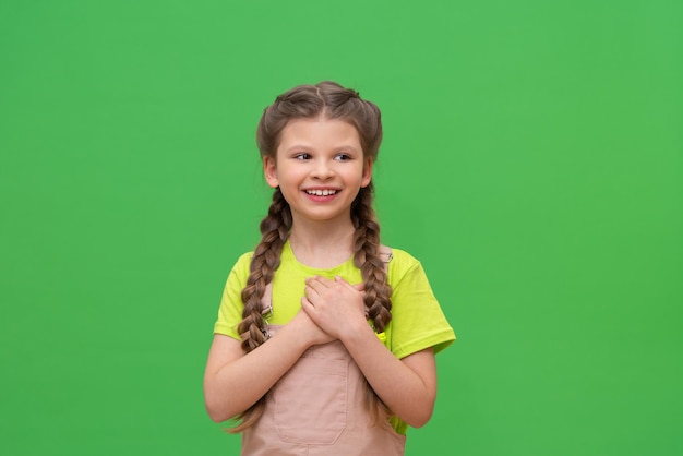 The girl put her hands to her heart and smiles. a little girl with pigtails on a green isolated background.