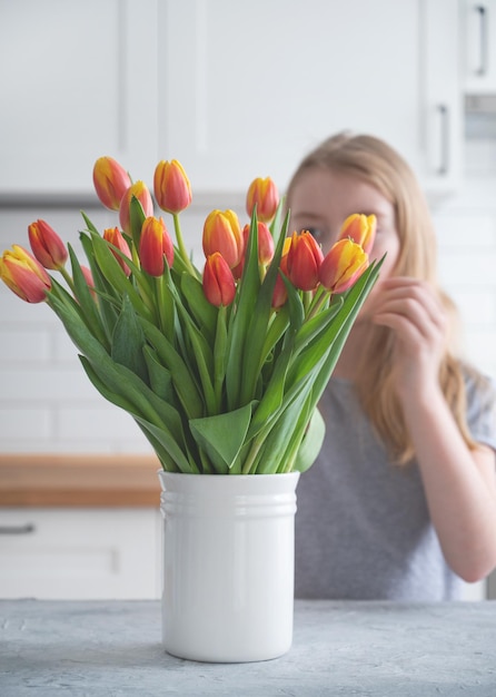 女の子は、白いキッチンを背景に木製のカウンタートップに白い花瓶に赤と黄色のチューリップの美しい花束を置きます 春の時間のコンセプト 色合いのイメージ