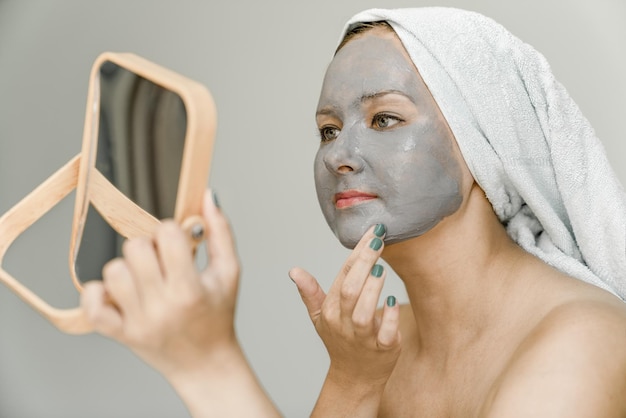 Girl puts gray cosmetic clay mask on face while looking in mirror her hair are wrapped in towel