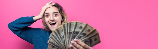 女の子は貯金箱に百ドル札を入れますピンクの背景の上の若い女の子