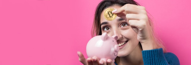 女の子は貯金箱を保持しているピンクの背景の上に物理的なBitcoinYoungの女の子を入れます