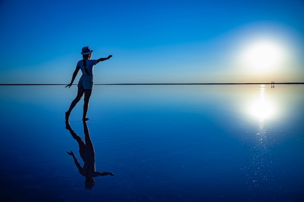 A girl reaches out with her hands to the setting sun on the mirror surface of a salt lake