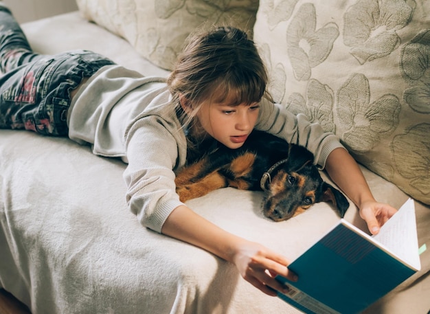 Девушка читает книгу, лежа на диване с собакой.