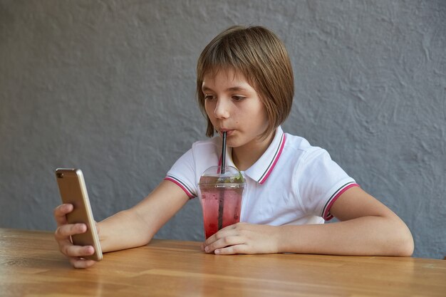 カフェのテーブルで赤い氷のカクテルを飲む電話で情報を読んでいる女の子